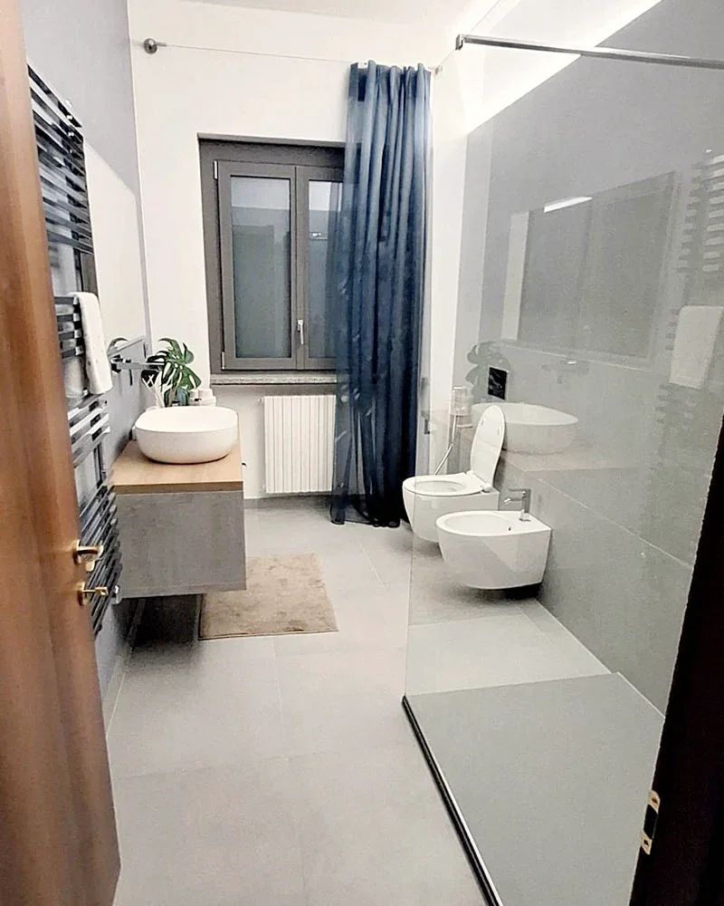 Lo stesso bagno dopo i lavori: doccia in vetro, rivestimenti chiari e sanitari sospesi